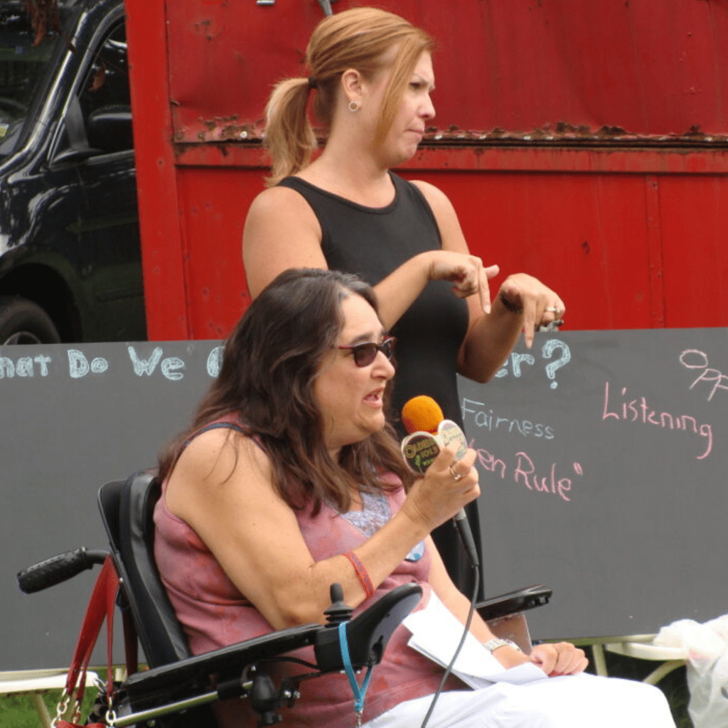 Fran, a white woman with long grey hair, and the founder of ATI, is sitting in her wheelchair and speaking into the microphone with an ASL interpreter behind her.